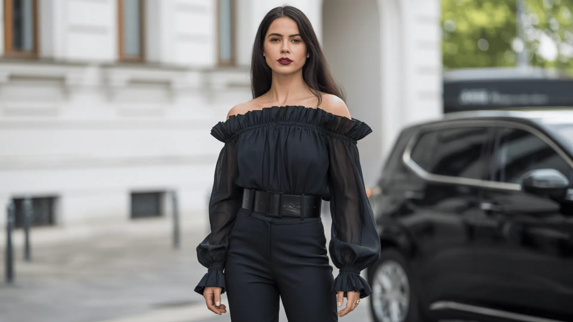 Off shoulder black blouse with sheer sleeves paired with high-waisted trousers in a city setting