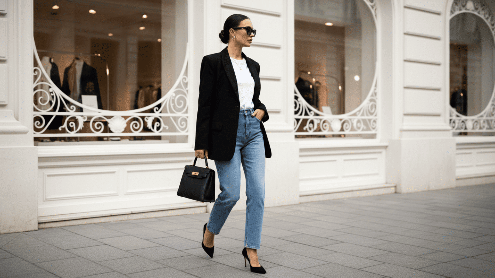Woman in casual chic outfit wearing a black blazer, white top, blue jeans, and black heels, walking with a structured handbag