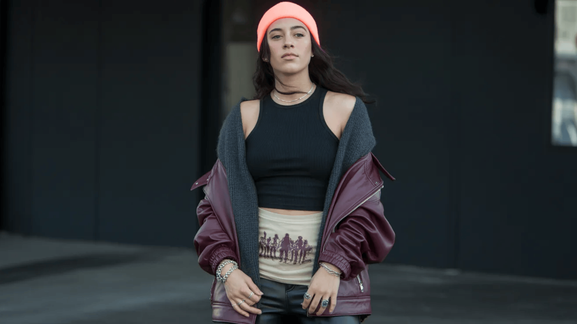 Indie sleaze look featuring a black crop top, leather jacket, dark layers, and a bold beanie accent against a moody urban backdrop