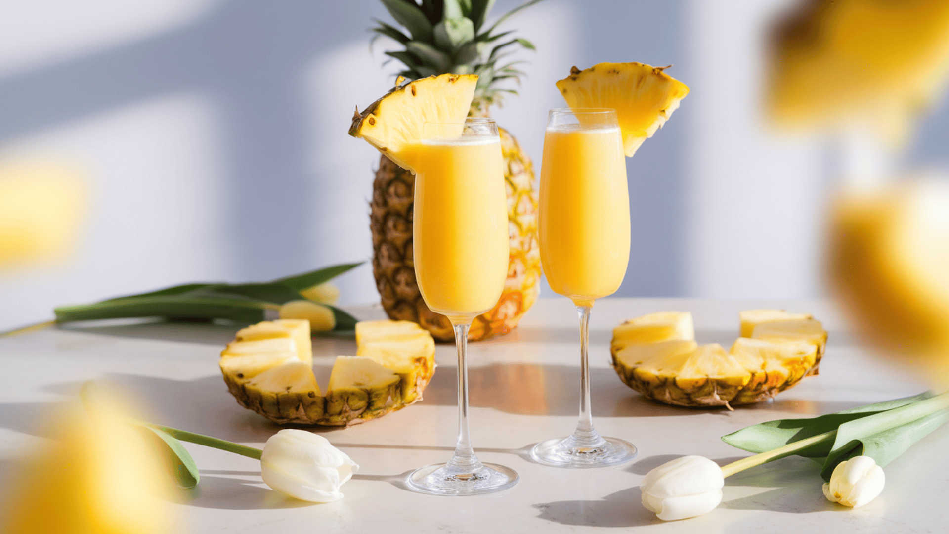 Two champagne flutes of pineapple mimosas garnished with pineapple wedges, surrounded by sliced pineapple and white tulips