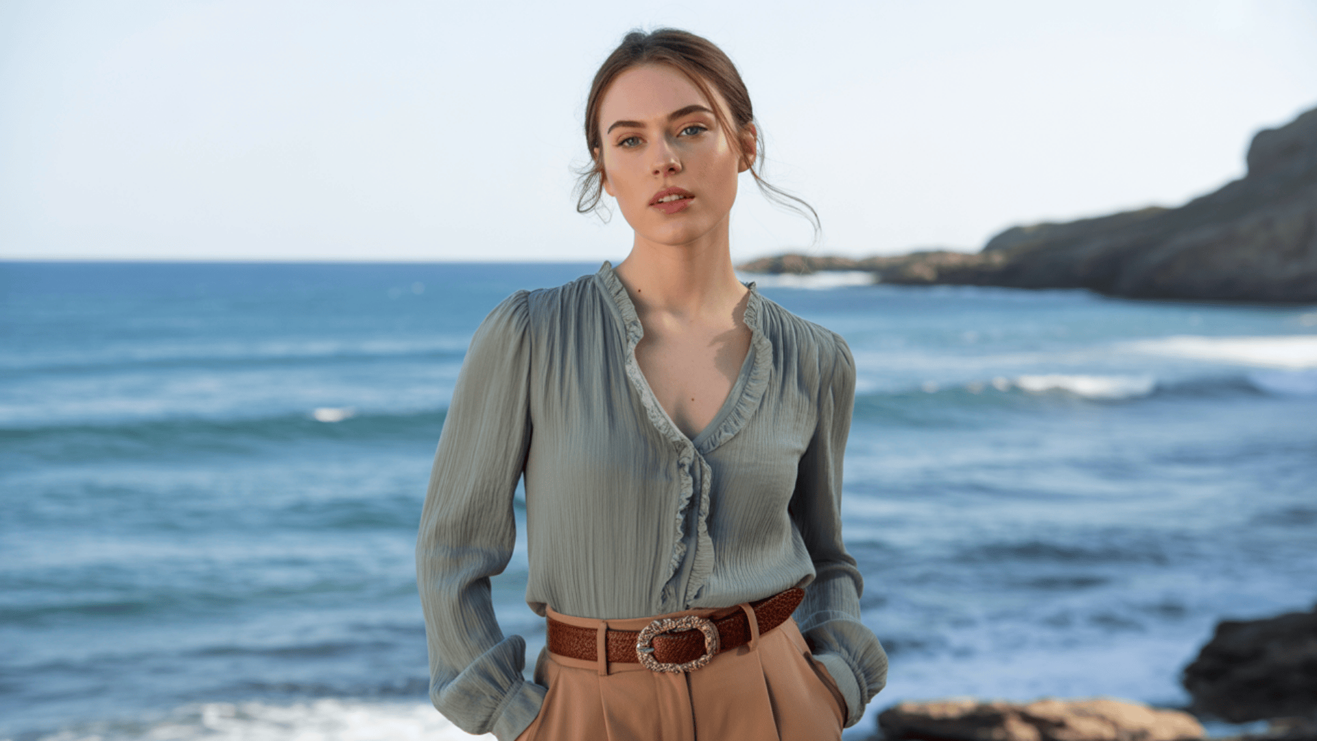 Woman wearing a soft ruffled blouse with long sleeves styled with a brown belt by the seaside.