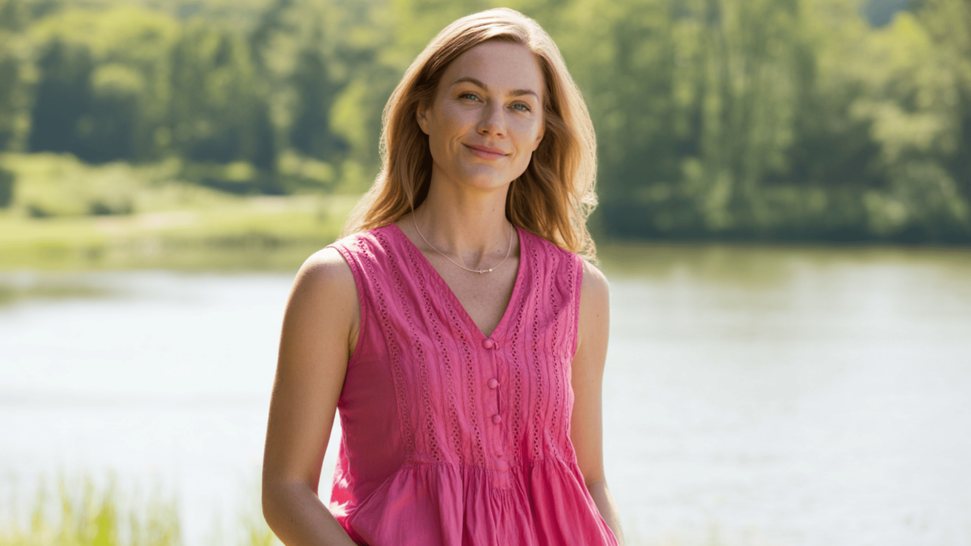 Woman wearing a pink sleeveless blouse standing outdoors near a lake with greenery in the background