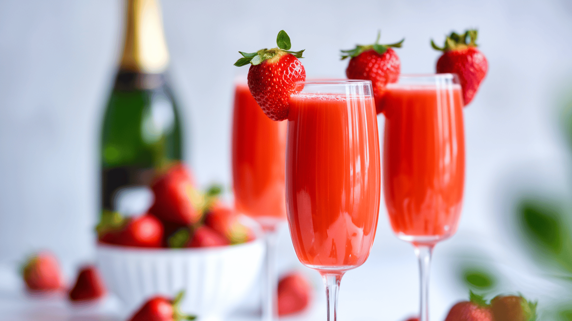 Three champagne flutes filled with strawberry mimosas, garnished with fresh strawberries, with a champagne bottle and bowl of strawberries behind