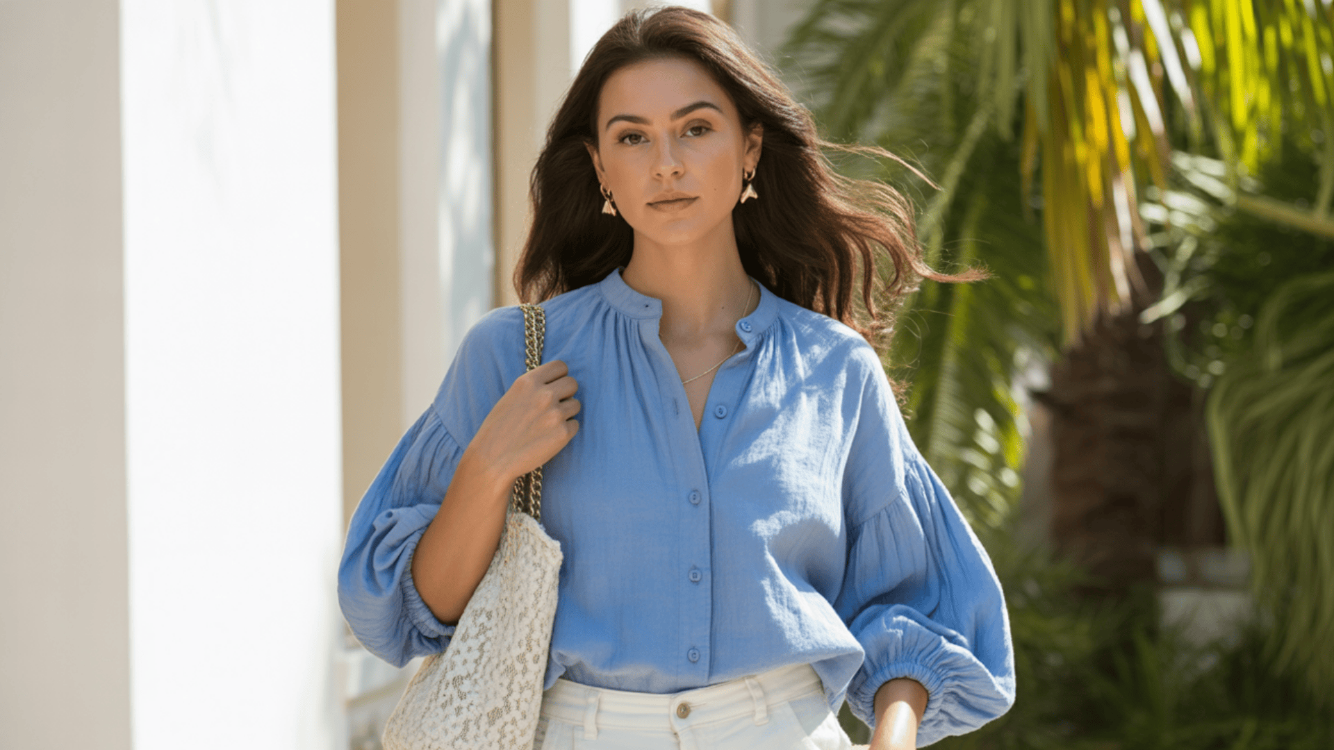 Woman wearing a lightweight blue blouse styled like a shirt, paired with white trousers and a woven tote outdoors