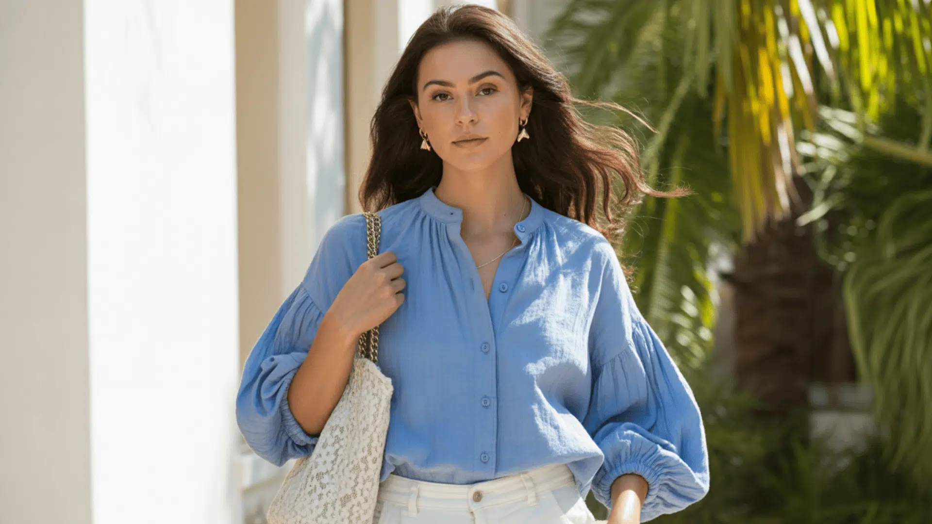 Woman wearing a lightweight blue blouse styled like a shirt, paired with white trousers and a woven tote outdoors