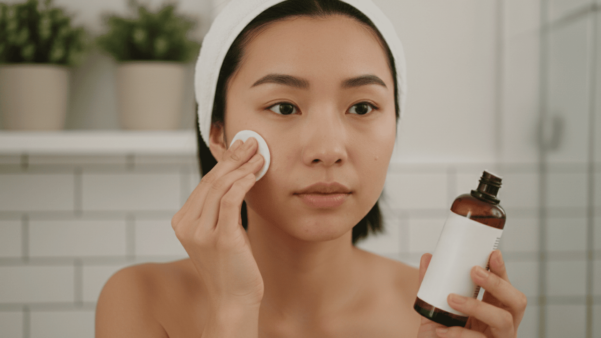 Woman applying facial toner with a cotton pad while holding a brown glass toner bottle in a bright bathroom