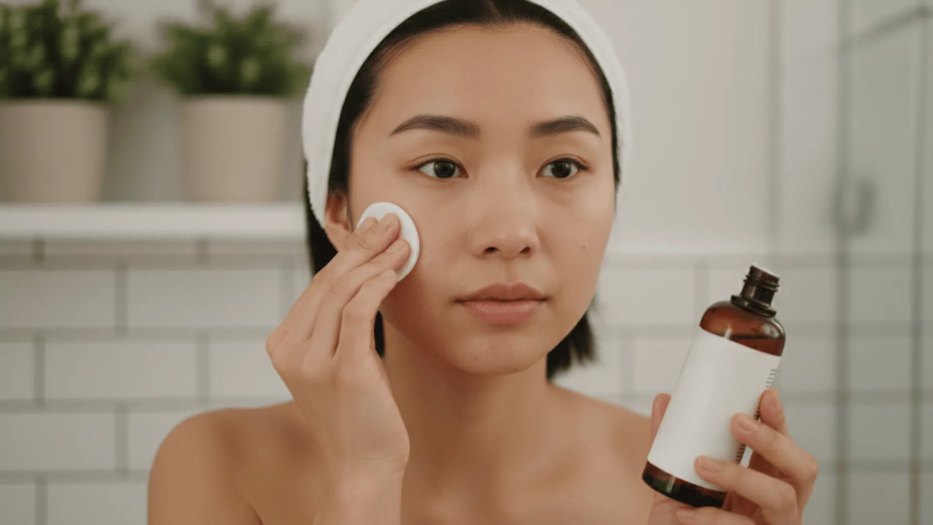 Woman applying facial toner with a cotton pad while holding a brown glass toner bottle in a bright bathroom