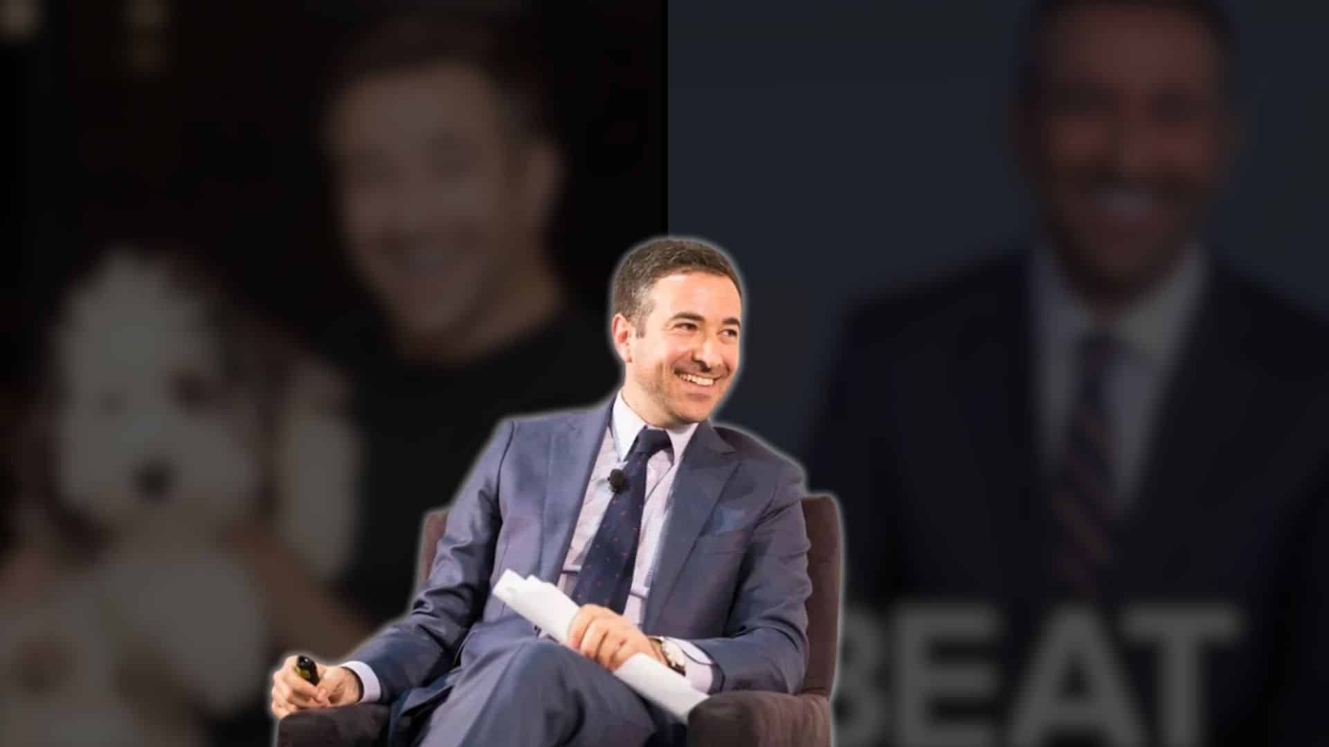 ari melber sitting on stage in a suit smiling during a public talk or event