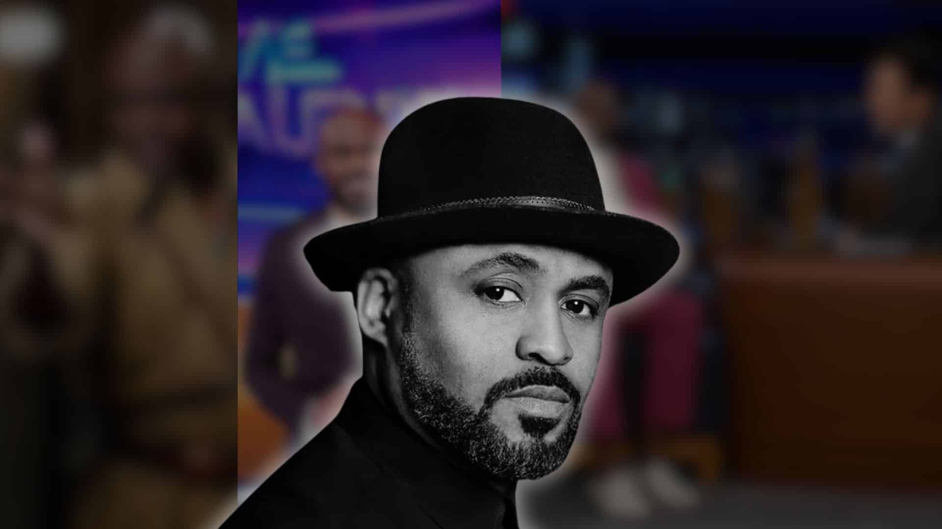 Close-up portrait of Wayne Brady wearing a black hat, with a blurred TV studio background
