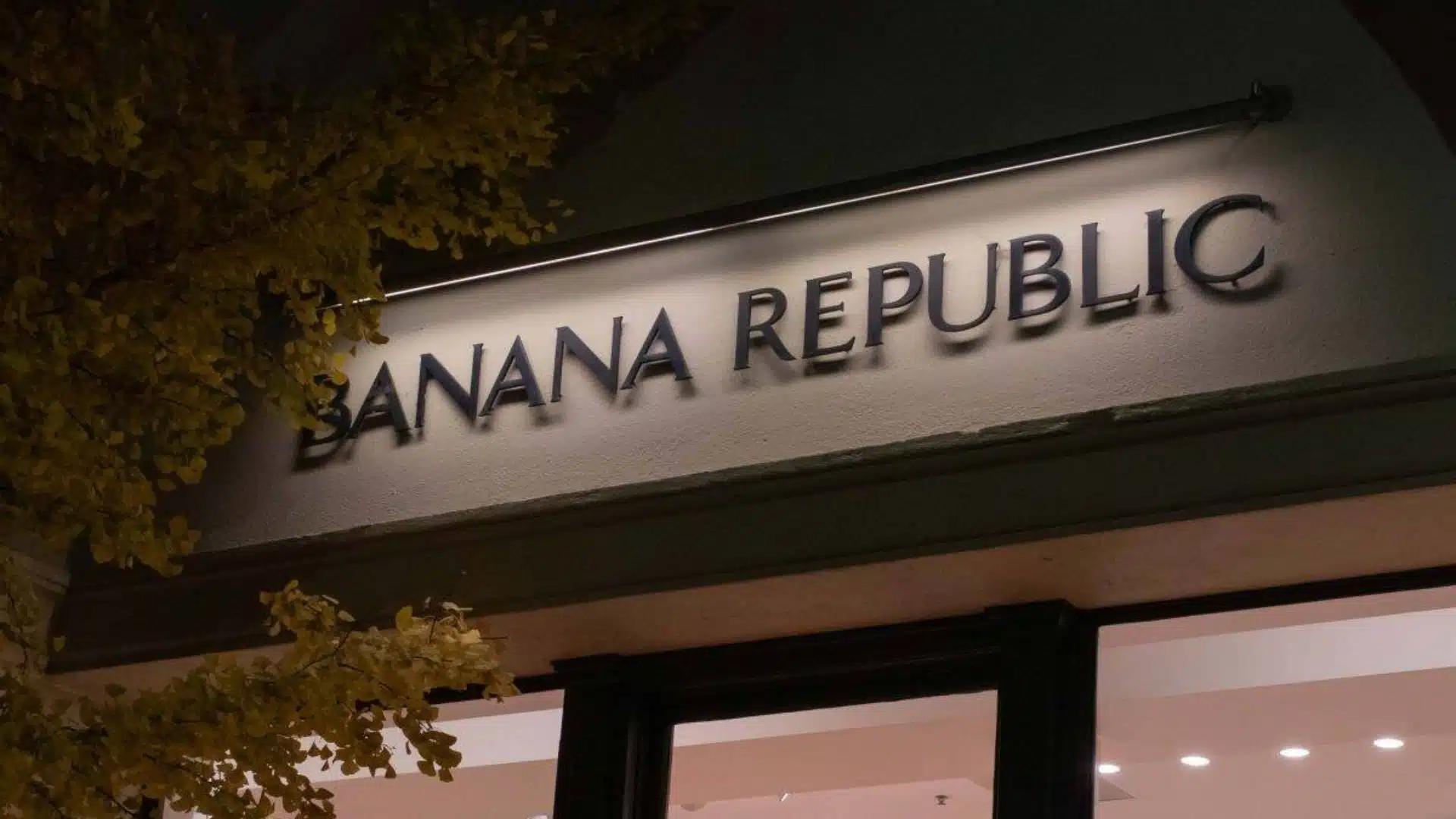 Banana Republic store sign illuminated at night with tree leaves framing storefront