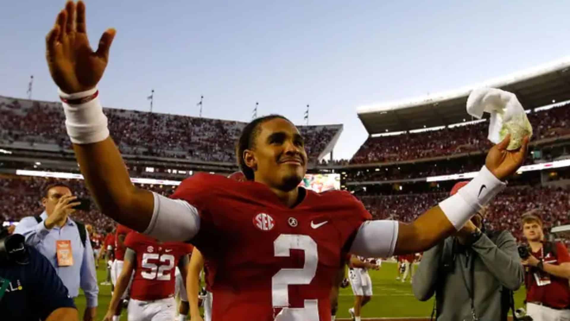 Jalen Hurts celebrates on the field in his Alabama Crimson Tide uniform, raising both arms as fans and teammates surround him after the game.