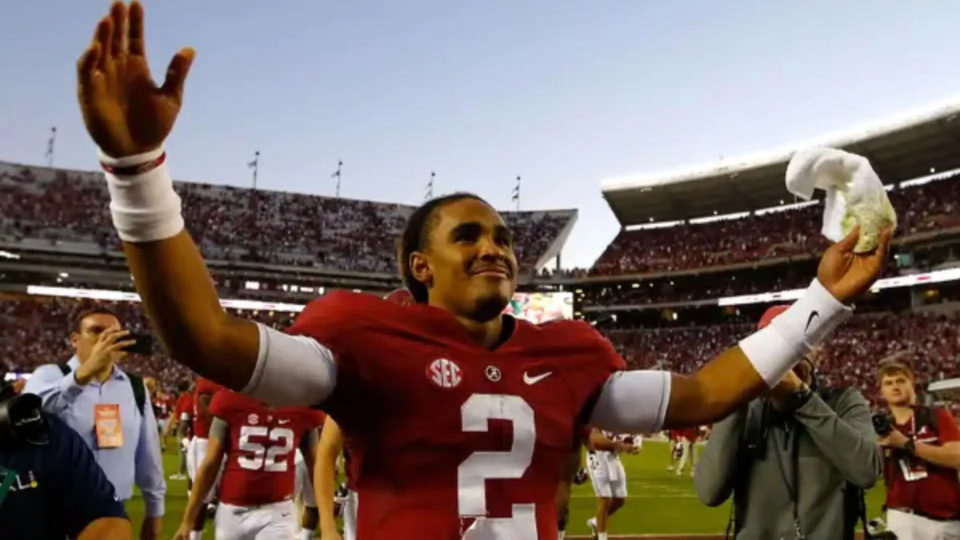 Jalen Hurts celebrates on the field in his Alabama Crimson Tide uniform, raising both arms as fans and teammates surround him after the game.