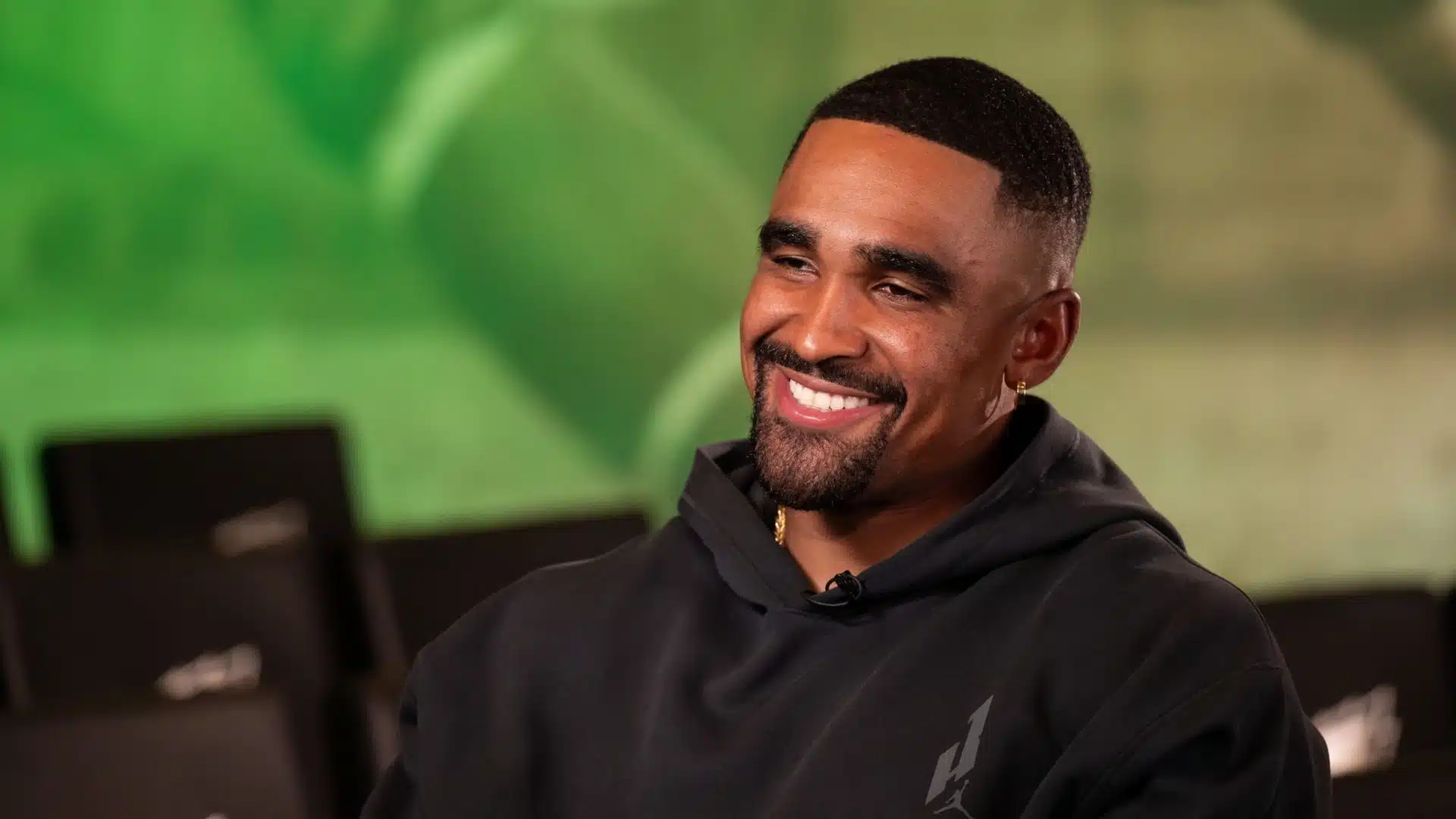 Jalen Hurts smiles during an interview while wearing a black hoodie, seated against a softly blurred green background.