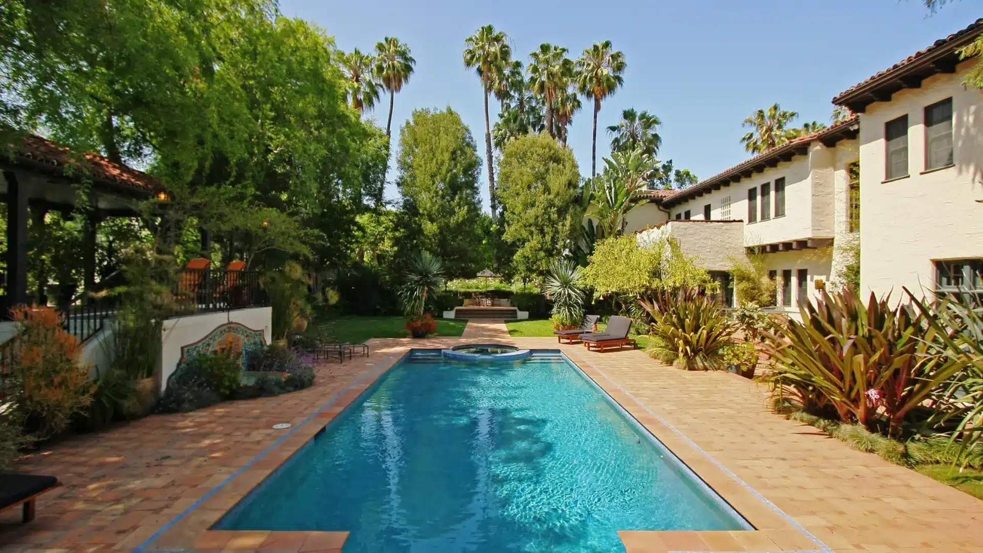 Luxury property exterior featuring a large swimming pool, landscaped garden, palm trees, and a Mediterranean-style home.
