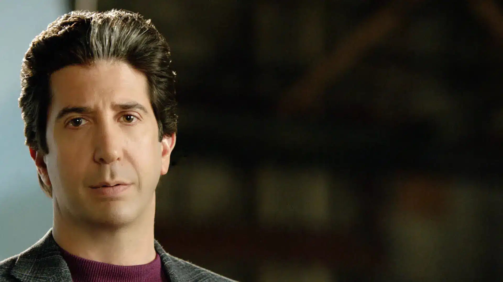 David Schwimmer in a serious close-up portrait as Robert Kardashian, shown against a blurred dark background in dramatic lighting.