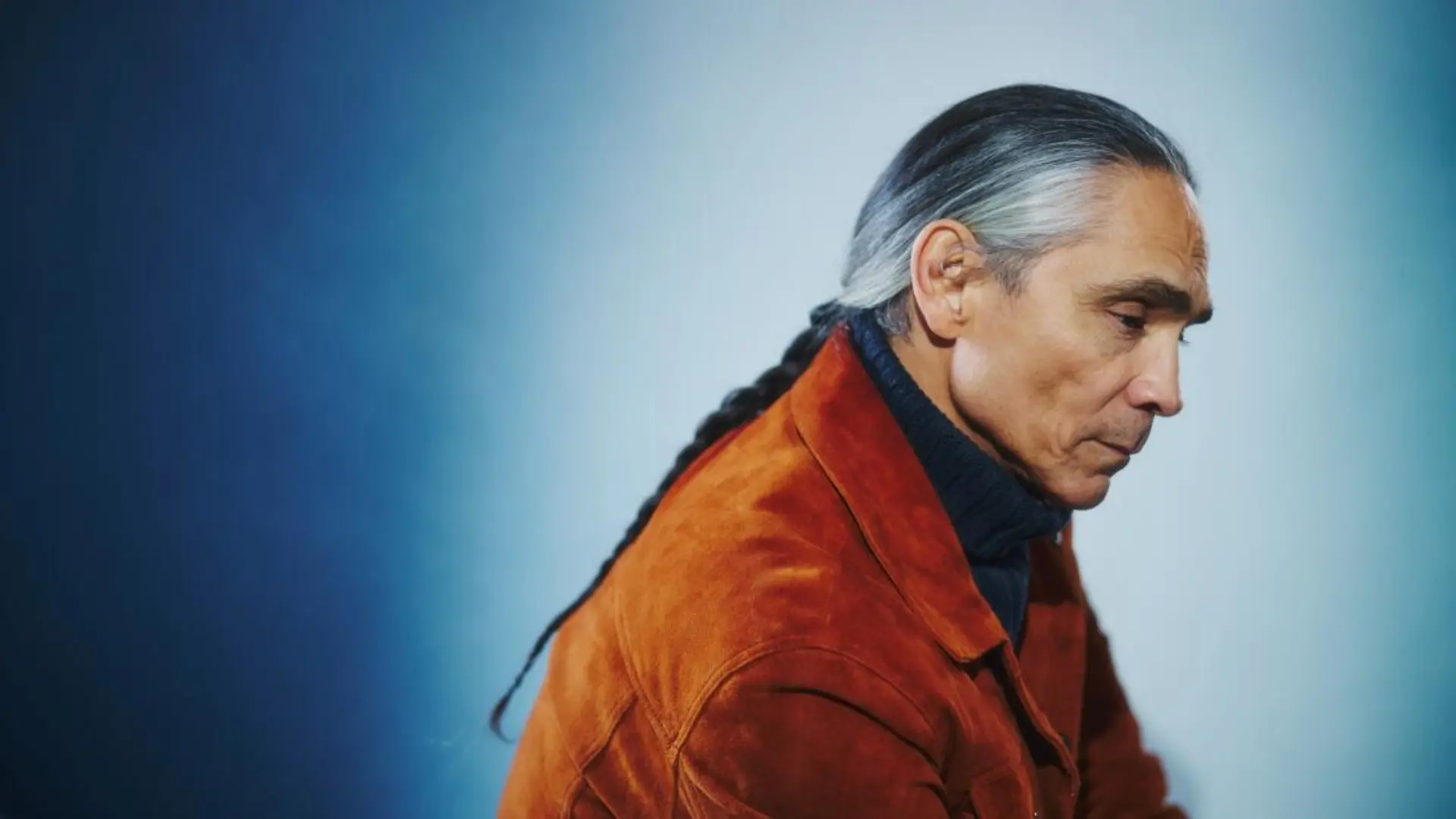 Side profile portrait of Zahn McClarnon with grey hair tied back, wearing an orange jacket against a blue gradient background.