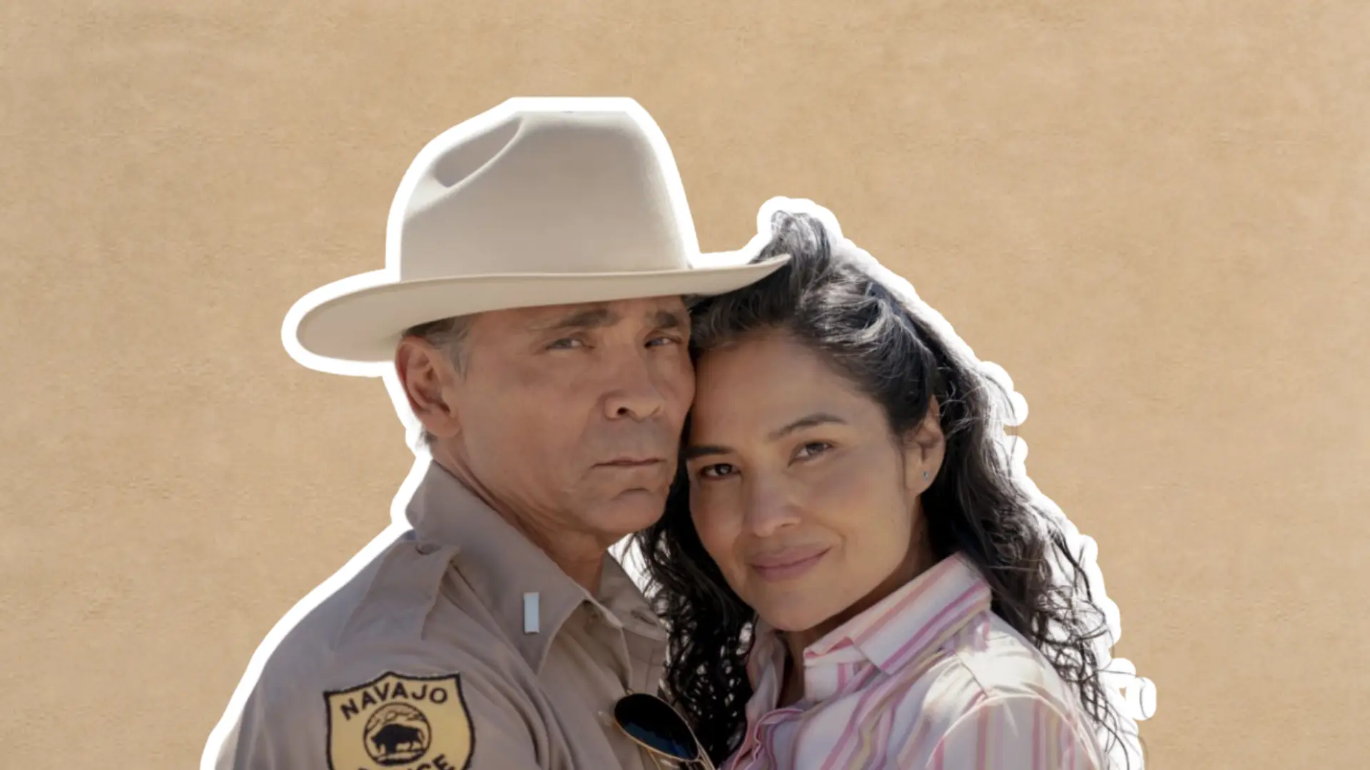 Zahn McClarnon in a police uniform hat standing close with a woman in a soft portrait-style pose.